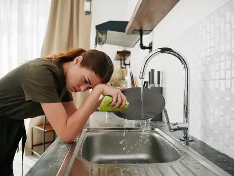 Уставшая женщина моет посуду на кухне под струей воды