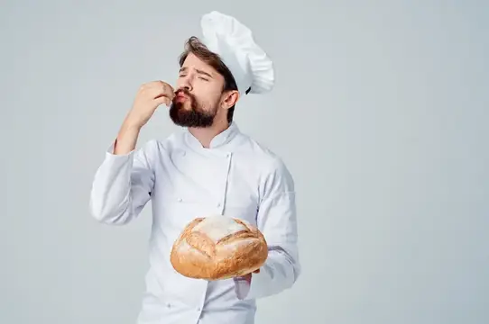 Бородатый пекарь в колпаке пробует хлеб на вкус