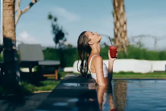 Молодая женщина с заплетёнными в косу волосами наслаждается освежающим красным напитком, отдыхая в современном открытом бассейне