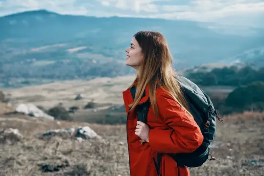 Улыбающаяся девушка с рюкзаком в красной куртке на фоне гор