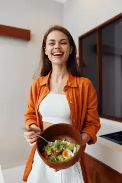 Улыбающаяся молодая женщина в оранжевой рубашке стоит на современной кухне, держа миску с разноцветным салатом