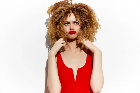 Молодая женщина с яркими вьющимися светлыми волосами, одетая в смелый красный наряд, демонстрирует игривое выражение лица на чистом белом фоне