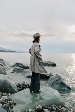 Женщина в плаще и кепке стоит на камнях у моря