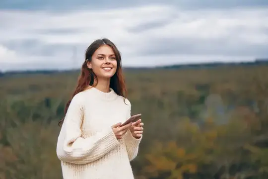 Улыбающаяся девушка в белом свитере с телефоном на фоне осеннего леса
