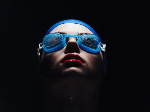 Пловчиха в синей шапочке и очках смотрит вверх на чёрном фоне