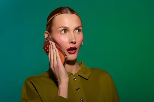 Удивлённая женщина с ярким макияжем слушает телефон на зелёном фоне