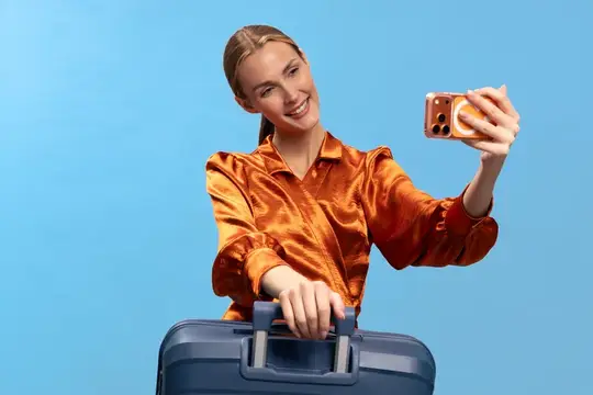 Улыбающаяся женщина в блестящей оранжевой рубашке делает селфи на телефон, держась за ручку чемодана, на фоне голубого фона