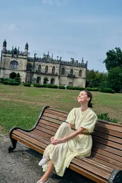 Молодая женщина в светлом платье отдыхает на скамейке у старинного замка