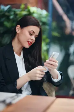 Улыбающаяся брюнетка в деловом костюме смотрит в смартфон в кафе