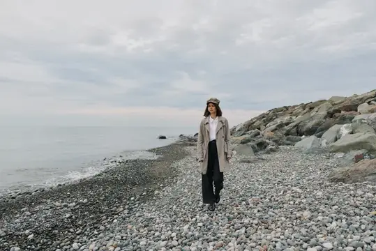 Молодая женщина в плаще и кепке гуляет по каменистому берегу моря