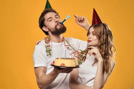 Счастливые мужчина и женщина в праздничных колпаках отмечают день рождения на жёлтом фоне, держа в руках торт