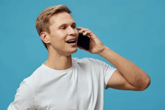 Улыбающийся светловолосый парень в белой футболке говорит по телефону на голубом фоне