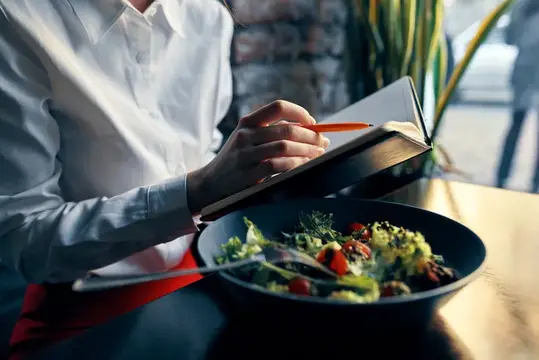 Блокнот с ручкой в руке и салат на тарелке в ресторане или кафе, присутствует надпись