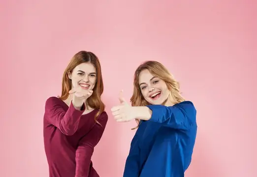 Две улыбающиеся девушки показывают большие пальцы на розовом фоне