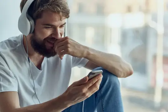 Бородатый мужчина в наушниках смеётся, глядя в смартфон