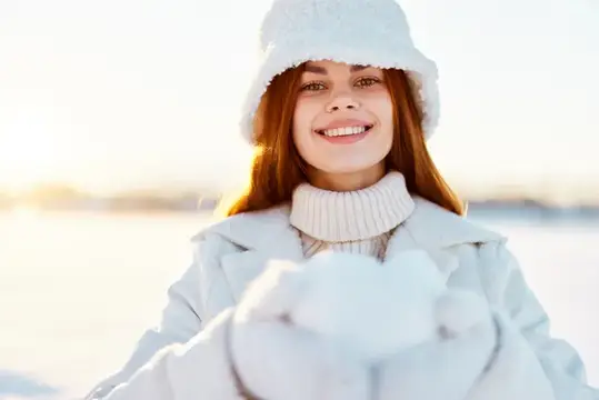 Улыбающаяся рыжеволосая девушка в белой шапке держит снег зимой