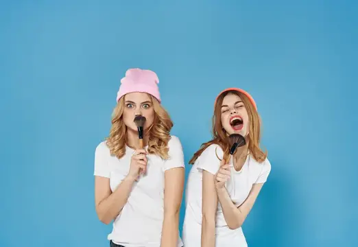 Две смешные девушки в шапках с кистями для макияжа на синем фоне