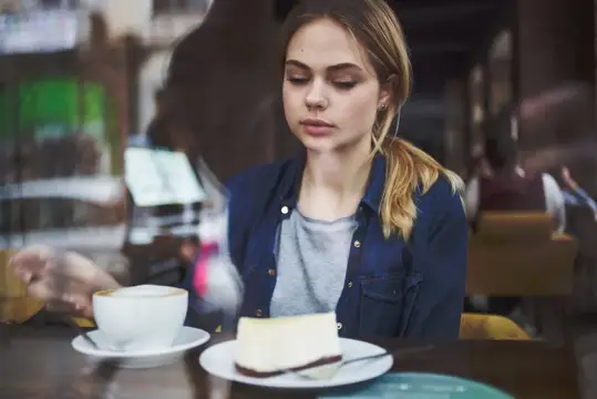 Задумчивая блондинка с чашкой кофе и куском торта у окна кафе