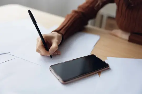 Рука в коричневом свитере рисует карандашом на листе бумаги рядом со смартфоном