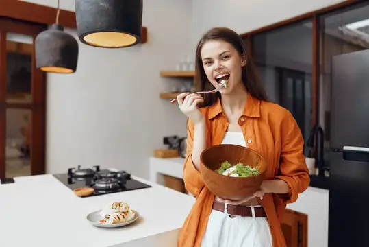Улыбающаяся девушка ест салат из деревянной миски на кухне
