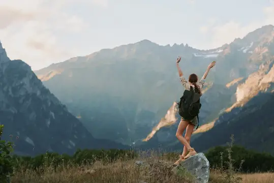 Одинокая женщина-туристка празднует достижение смотровой площадки, одетая в повседневную одежду, на заднем плане скалистые вершины и ясное небо, что передает свободу, уверенность и дух приключений…