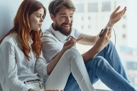 Рыжеволосая девушка и бородатый мужчина весело смотрят в смартфон у окна