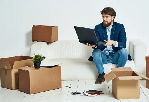 Мужчина сидит на диване с ноутбуком, офисными коробками с вещами, работает