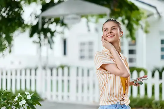 Улыбающаяся девушка в наушниках слушает музыку у белого дома