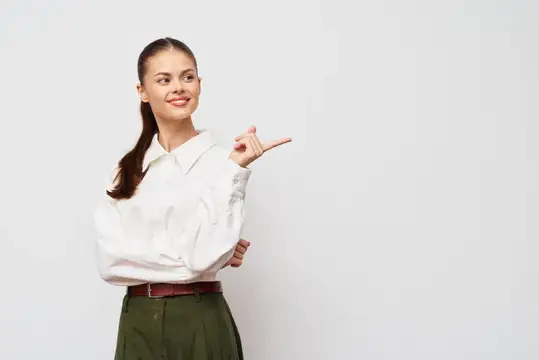 Уверенная бизнес-леди улыбается и делает указывающий жест правой рукой, одетая в белую блузку и зелёные брюки, позирует на минималистичном светло-сером фоне