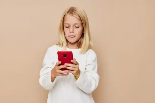 Белокурая девочка в белом свитере смотрит в красный смартфон на бежевом фоне