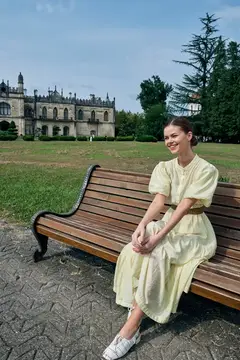 Улыбающаяся девушка в светлом платье сидит на скамейке в парке у замка
