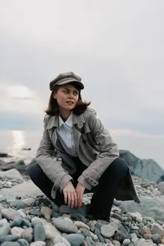Молодая женщина в кепке и плаще сидит на камнях у моря