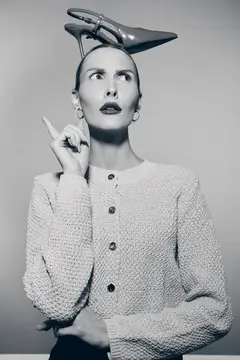 Черно-белое изображение светлокожей женщины-европейки с туфлей на высоком каблуке на голове, указывающей вверх и выражающей любопытство или задумчивость в студии
