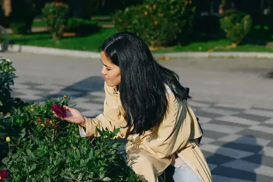 Темноволосая девушка в бежевом плаще трогает цветы на клумбе