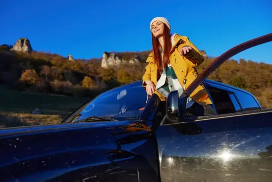 Рыжеволосая девушка в жёлтой куртке выглядывает из люка автомобиля на фоне осенних гор