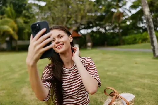Улыбающаяся темноволосая девушка делает селфи на лужайке в парке