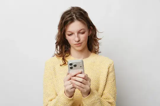 Женщина в жёлтом вязаном свитере держит смартфон обеими руками, сосредоточенно глядя на экран