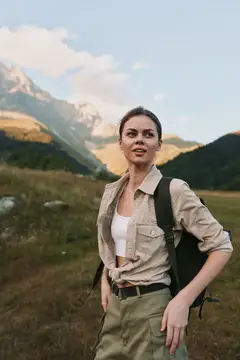 Уверенная женщина наслаждается приключением на свежем воздухе, одетая в повседневную экипировку с рюкзаком; она стоит среди травы и альпийских пейзажей в солнечной атмосфере, с естественными тонами…