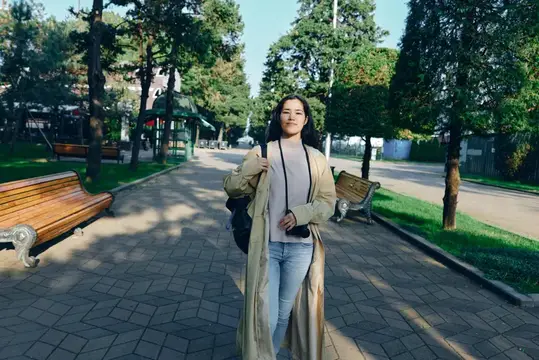 Женщина в бежевом пальто прогуливается по парковой дорожке под деревьями