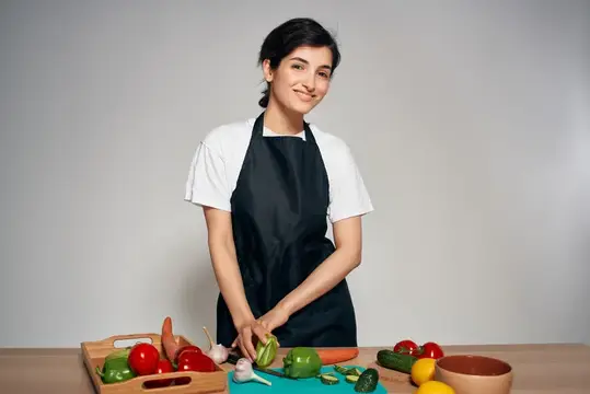Улыбчивая темноволосая женщина в фартуке режет овощи на кухне