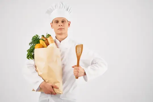 Молодой повар в колпаке с бумажным пакетом продуктов и деревянной лопаткой