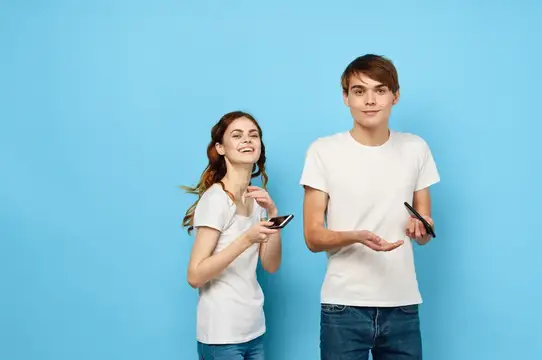 Весёлая молодая пара в белых футболках с телефонами в руках ведёт общение, демонстрируя определённый образ жизни