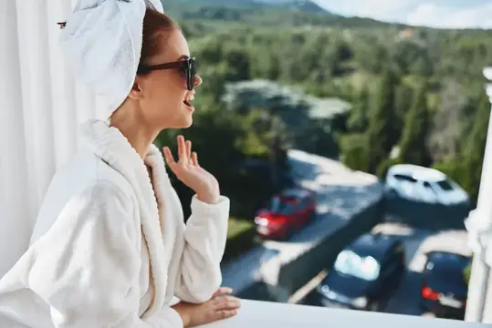 Улыбающаяся женщина в халате и полотенце на голове смотрит с балкона на горы