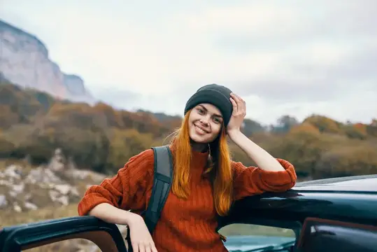 Женщина-туристка с рюкзаком стоит возле машины на природе, в горном путешествии