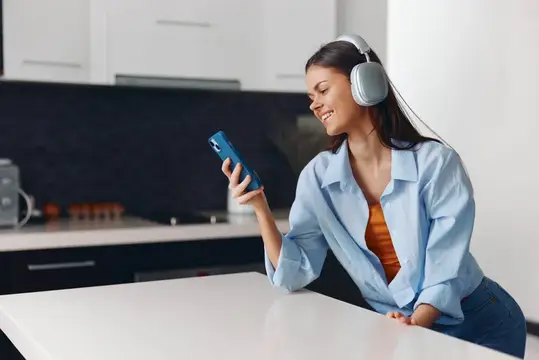 Женщина наслаждается музыкой в наушниках через телефон, сидя за кухонным столом