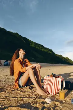 Женщина на пляже, песок, море, лето, отдых, полоски, солнечный свет, момент пляжного туризма: она сидит, скрестив ноги, на песке лежат полосатая сумка и сумочка, соломенная шляпа, шарф, мирная и…