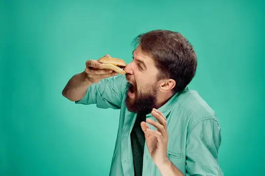 Бородатый мужчина с аппетитом кусает сэндвич на бирюзовом фоне
