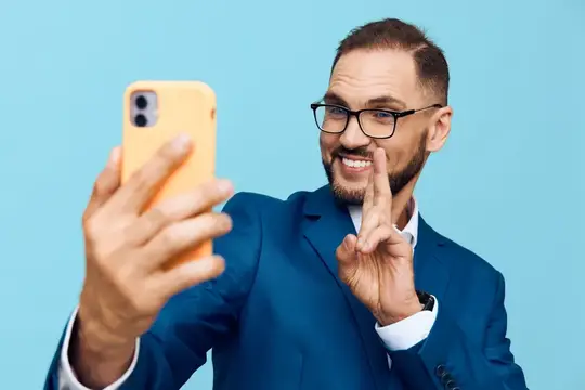 Улыбающийся бородатый мужчина в очках делает селфи на жёлтый смартфон