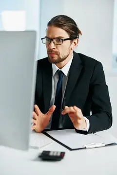 Мужчина в очках и костюме жестикулирует перед монитором в офисе