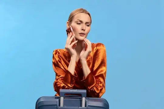 Блондинка в оранжевой блузе говорит по телефону у чемодана
